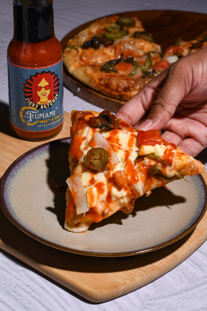 Person holding a slice of pizza drizzled with Fumami sweet and spicy fermented Fiery Ketchup on a wooden board.