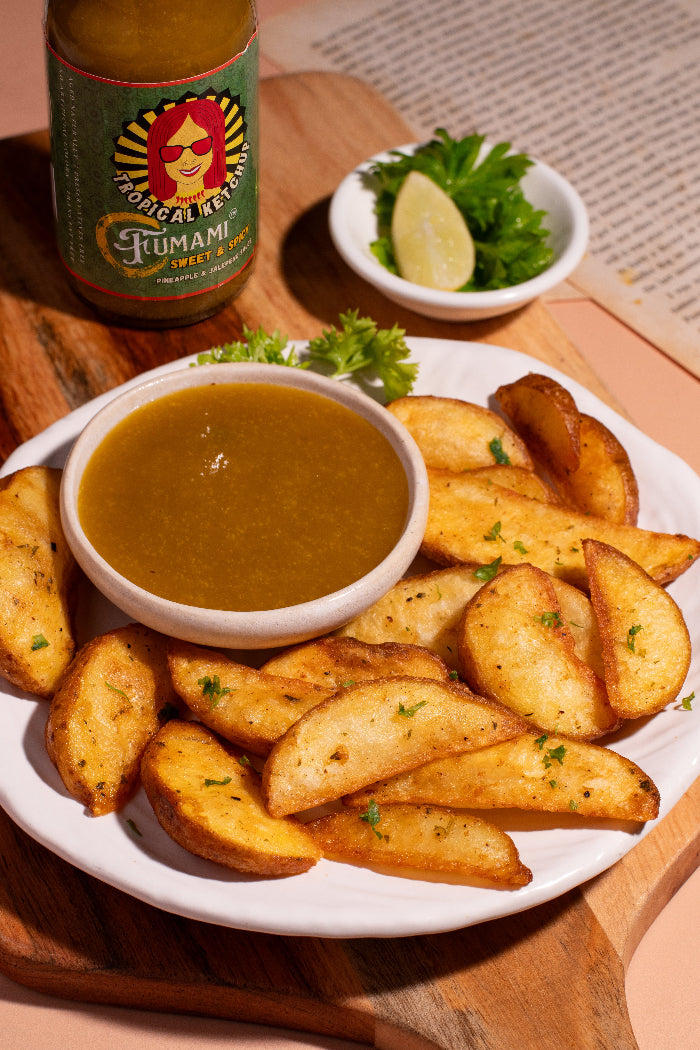 Platter of potato wedges with a bowl of Fumami sweet and spicy fermented tropical ketchup sauce and a bottle of fumami on a wooden surface.